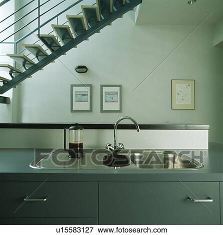 Stock Photo - Coffeepot beside sink in blue-grey fitted unit below staircase in modern kitchen. Fotosearch