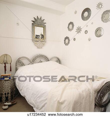 Stock Image - Collection of tin mirrors on wall above carved bed with white duvet in small white bedroom. Fotosearch