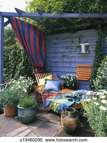 Stock Image - Colourful rug and cushions on chaiselongue below painted blue pergola with striped curtain in summer garden. Fotosearch