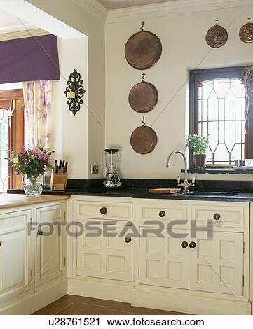 Stock Image - Copper pans on wall above cream fitted units in traditional country kitchen. Fotosearch