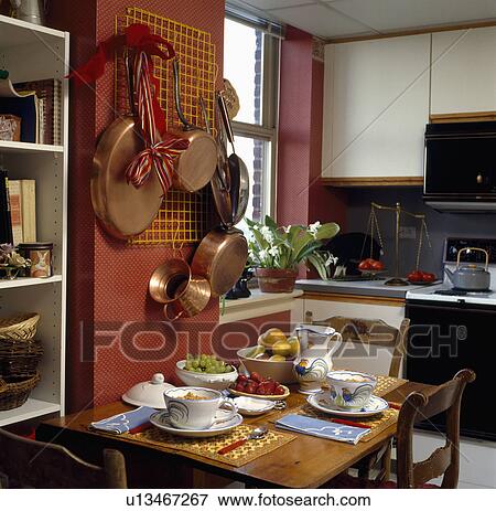 Stock Photo - Copper pans on wall above small wooden table with pottery crockery in small townhouse kitchen. Fotosearch