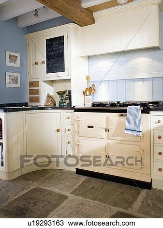 Stock Image - Cream Aga oven in pastel blue country kitchen with flagstone floor and cream fitted units. Fotosearch