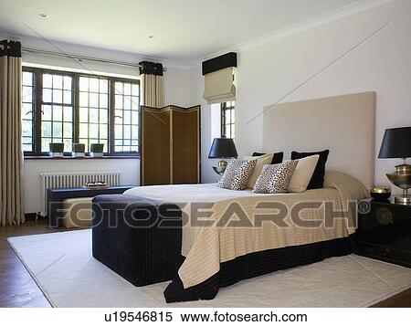 Stock Photography - Cream bedcover and animal-print cushions on bed with large beige suede headboard in modern country bedroom. Fotosearch