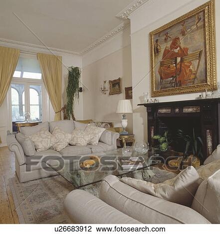 Stock Image - Cream sofas and modern glass table in traditional living room with large picture above fireplace. Fotosearch