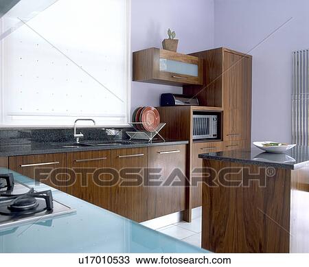 Stock Image - Dark brown fitted units in modern mauve kitchen with white blind on window. Fotosearch