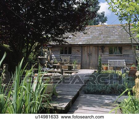 Stock Image - Decking path across stream to single-storey wooden country cottage. Fotosearch