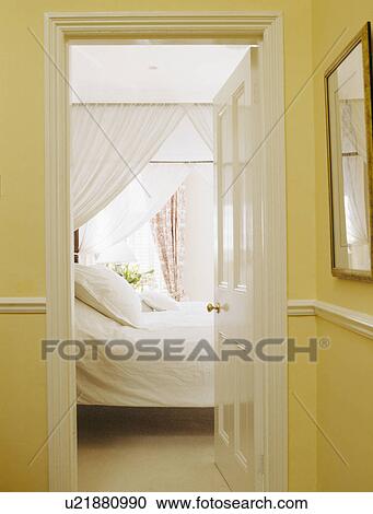Stock Image - Door open to bed with white drapes and bedlinen. Fotosearch