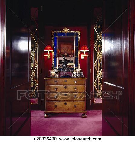 Stock Image - Double mahogany doors open to dressing room with ornate blue+gold framed mirror above antique chest-of-drawers. Fotosearch