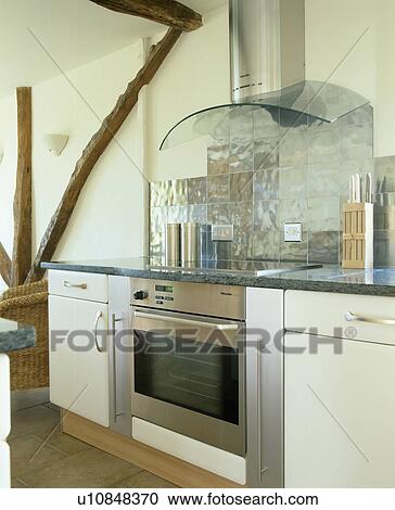Stock Image - Extractor and metallic wall-tiles above oven in modern white fitted unit in beamed cottage kitchen. Fotosearch