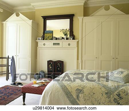 Picture - Fitted cream wardrobes in alcoves on either side of fireplace in pale yellow bedroom. Fotosearch