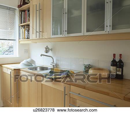 Stock Photo - Fitted cupboard with glazed doors above sink set into pale wood units. Fotosearch