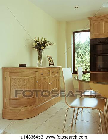 Stock Image - Fitted sideboard in modern cream kitchen with cream ceramic tiled floor with pale wood chairs at breakfast bar. Fotosearch