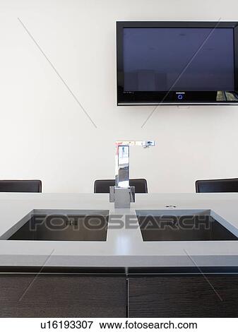 Stock Photo - Flatscreen television on wall above stainless-steel tap and underset double sink in grey worktop. Fotosearch
