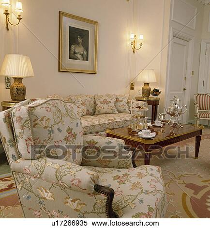 Stock Photography - Floral armchair and sofa in townhouse drawing room with silver teaset on coffee table. Fotosearch