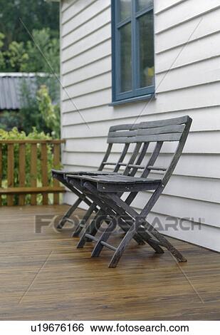 Stock Photograph - Folding black wooden chairs on decking in front of white ship-lap house. Fotosearch