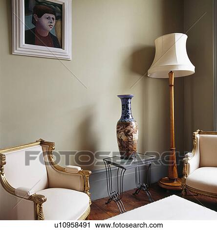 Stock Image - Gilt-framed French armchairs and tall vase on glass-topped metal table in living room with cream standard lamp. Fotosearch