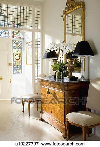 Stock Photo - Gilt mirror above antique chest-of-drawers with black lamps in traditional white hall with limestone floor tiles. Fotosearch