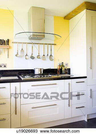 Stock Image - Glass and stainless steel extractor and steel utensils on wall-rack above hob in modern white fitted kitchen unit. Fotosearch