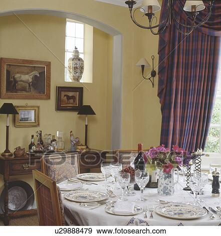 Stock Photo - Glasses and plate settings on white linen cloth on table in traditional dining room with checked swagged curtains. Fotosearch