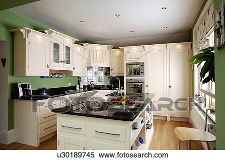 Stock Photography - Granite-topped island unit in green kitchen with white fitted units. Fotosearch