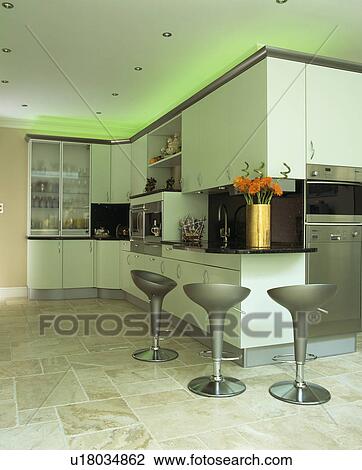 Stock Image - Green halogen lighting above fitted white cupboards in modern kitchen with Bomba stools and limestone flooring. Fotosearch