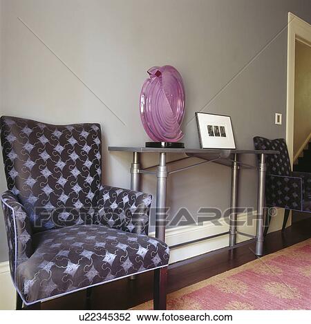 Stock Image - Grey & black upholstered armchair beside glass-topped steel console table with large mauve glass vase in grey hall. Fotosearch