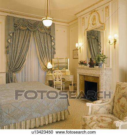 Stock Image - Grey swagged and draped curtains on window in townhouse bedroom with mirror above marble fireplace. Fotosearch