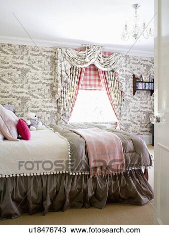 Stock Image - Grey Toile-de-Jouy wallpaper and curtains in country bedroom with checked blanket and grey quilt on bed. Fotosearch