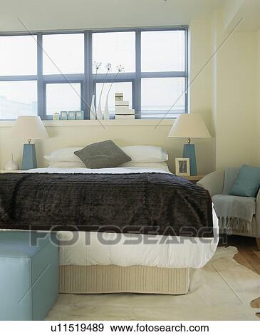 Stock Photo - Grey velvet throw on bed with white bedlinen below large window in modern white bedroom. Fotosearch