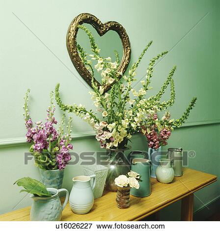 Stock Photo - Heart-shaped mirror on pastel turquoise wall above pink and white foxgloves in vases and collection of green jugs. Fotosearch
