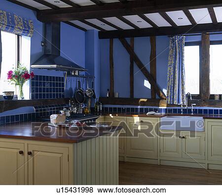 Stock Photo - Iroko worktops on cream fitted units in blue country kitchen with beamed ceiling. Fotosearch