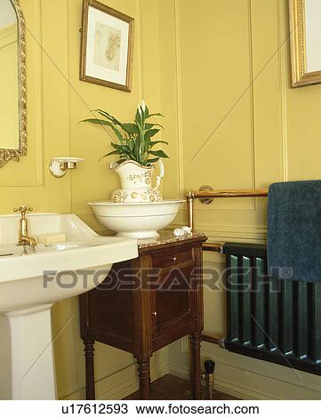 Stock Image - Jug and bowl on Edwardian cabinet beside white pedestal basin in yellow panelled bathroom. Fotosearch