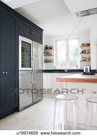 Stock Photo - Kartell perspex stools at breakfast bar in modern white kitchen with large stainless steel fridge-freezer. Fotosearch