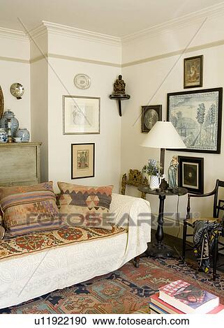 Stock Image - Kelim cushions and cream throw on sofa in living room with pictures on wall behind white lamp on small table. Fotosearch