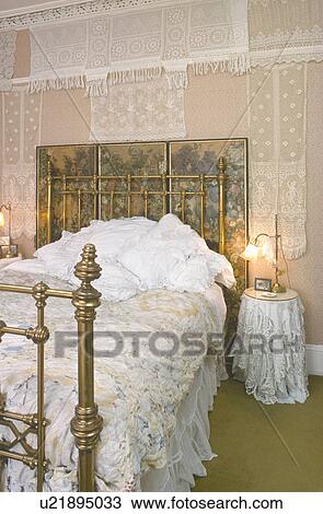 Stock Image - Lace covered wall behind antique brass bed in Victorian bedroom with lace bedcover. Fotosearch