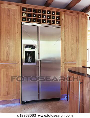 Stock Image - Large American-style stainless steel fridge freezer fitted between wood storage cupboards in modern kitchen. Fotosearch