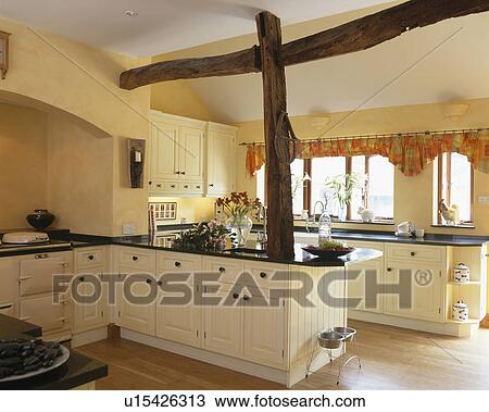 Stock Image - Large beams in traditional yellow country kitchen with cream fitted units. Fotosearch
