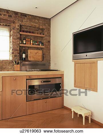 Stock Image - Large flatscreen television fitted into wall in modern kitchen with pale wood fitted units and stainless steel oven. Fotosearch