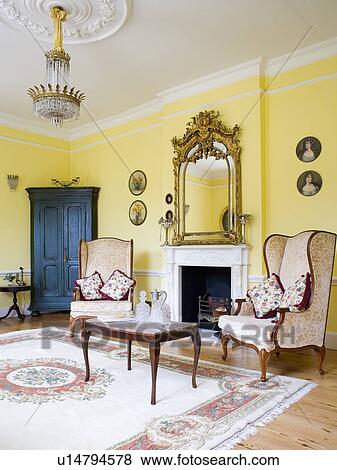 Stock Photo - Large gilt antique mirror above fireplace in yellow living room with cream wing-chairs and patterned rug. Fotosearch