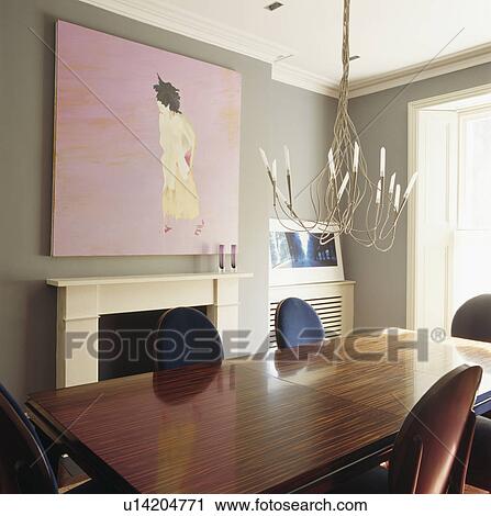 Stock Image - Large modern picture above fireplace in dining room with wirework chandelier above polished wooden table. Fotosearch