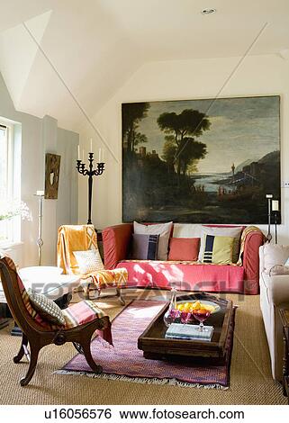 Large oil painting above red sofa piled with cushions in modern living room with antique Indian planters chair and table View Large Photo Image Stock Photograph - Large oil painting above red sofa piled with cushions in modern living room with antique Indian planters chair and table. Fotosearch