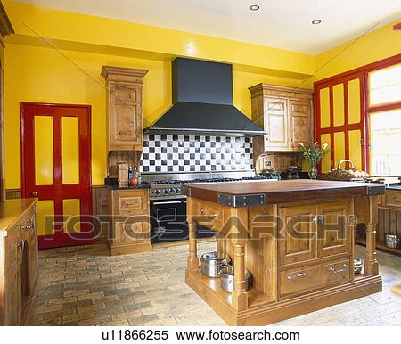 Stock Photography - Large wooden island unit in yellow kitchen with red paintwork and brick flooring. Fotosearch