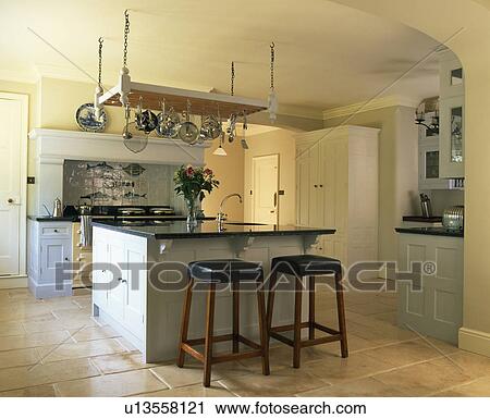 Stock Image - Leather and wood stools at breakfast bar on island unit in country kitchen with limestone flooring. Fotosearch