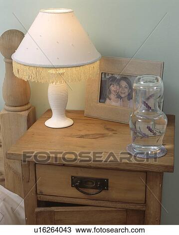 Stock Image - Lighted lamp and water carafe on pine bedside table. Fotosearch
