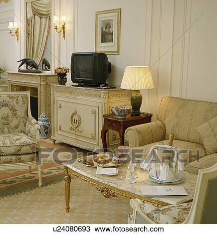 Stock Image - Lighted lamp on small table beside beige armchair in traditional townhouse living room. Fotosearch