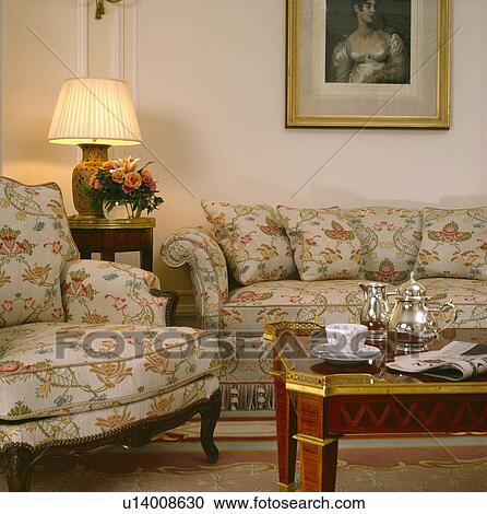 Stock Image - Lighted lamp on small table between floral patterned sofa and armchair in living room with silver teaset on coffee table. Fotosearch