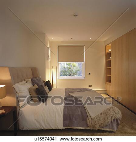 Stock Image - Lighted lamps and neutral bedlinen in townhouse bedroom with cream blind. Fotosearch