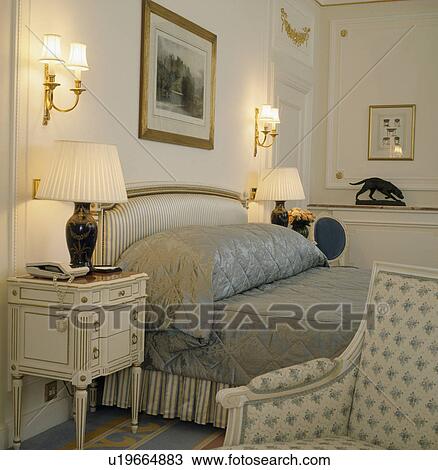 Lighted lamps on bedside tables on either side of upholstered bed with grey bedlinen in townhouse bedroom View Large Photo Image Stock Image - Lighted lamps on bedside tables on either side of upholstered bed with grey bedlinen in townhouse bedroom. Fotosearch