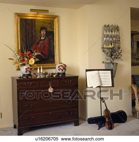 Stock Photography - Lighting above gilt-framed oil portrait on wall above antique chest of drawers beside music stand and violin. Fotosearch