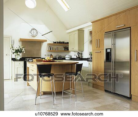 Stock Photo - Metal stools at breakfast bar on island unit in modern kitchen with fitted stainless steel American-style fridge-freezer. Fotosearch
